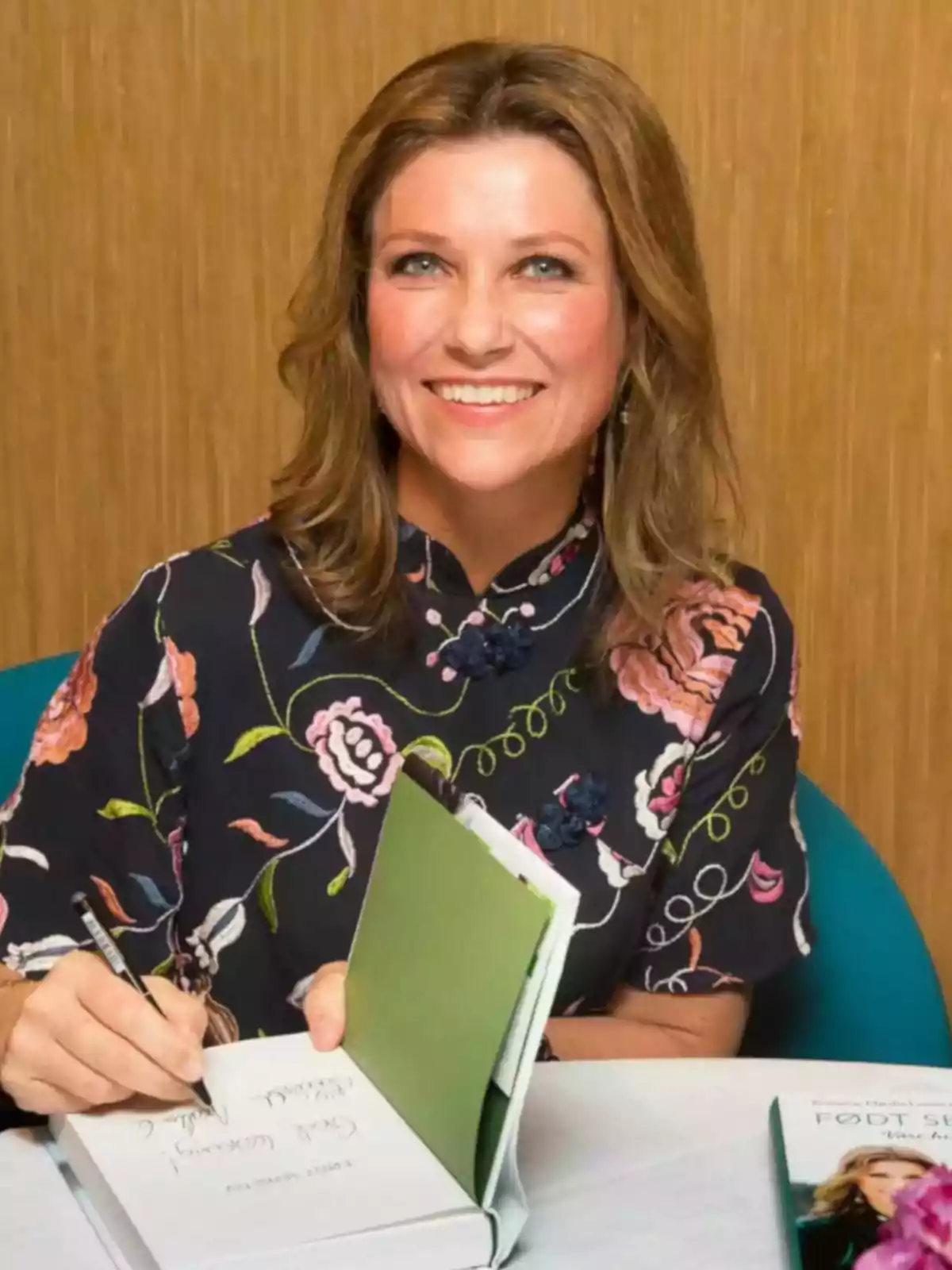 Dona somrient amb cabell castany clar signant un llibre mentre està asseguda a una taula, porta una brusa fosca amb estampat floral Dona somrient amb cabell castany clar signant un llibre mentre està asseguda a una taula, porta una brusa fosca amb estampat floral