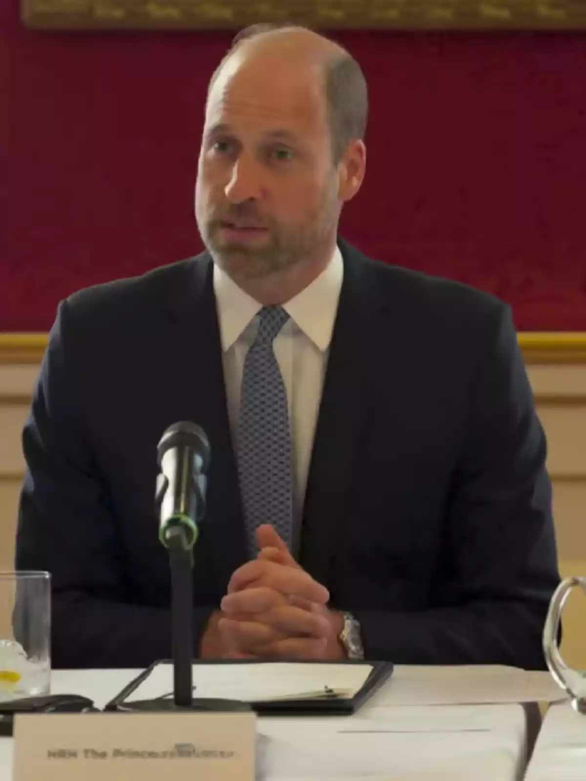 Hombre con traje y corbata azul hablando frente a un micrófono en una mesa de conferencia Hombre con traje y corbata azul hablando frente a un micrófono en una mesa de conferencia