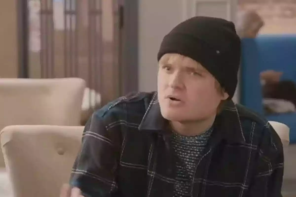 Joven con gorro negro y camisa de cuadros sentado en una cafetería con expresión de sorpresa Joven con gorro negro y camisa de cuadros sentado en una cafetería con expresión de sorpresa