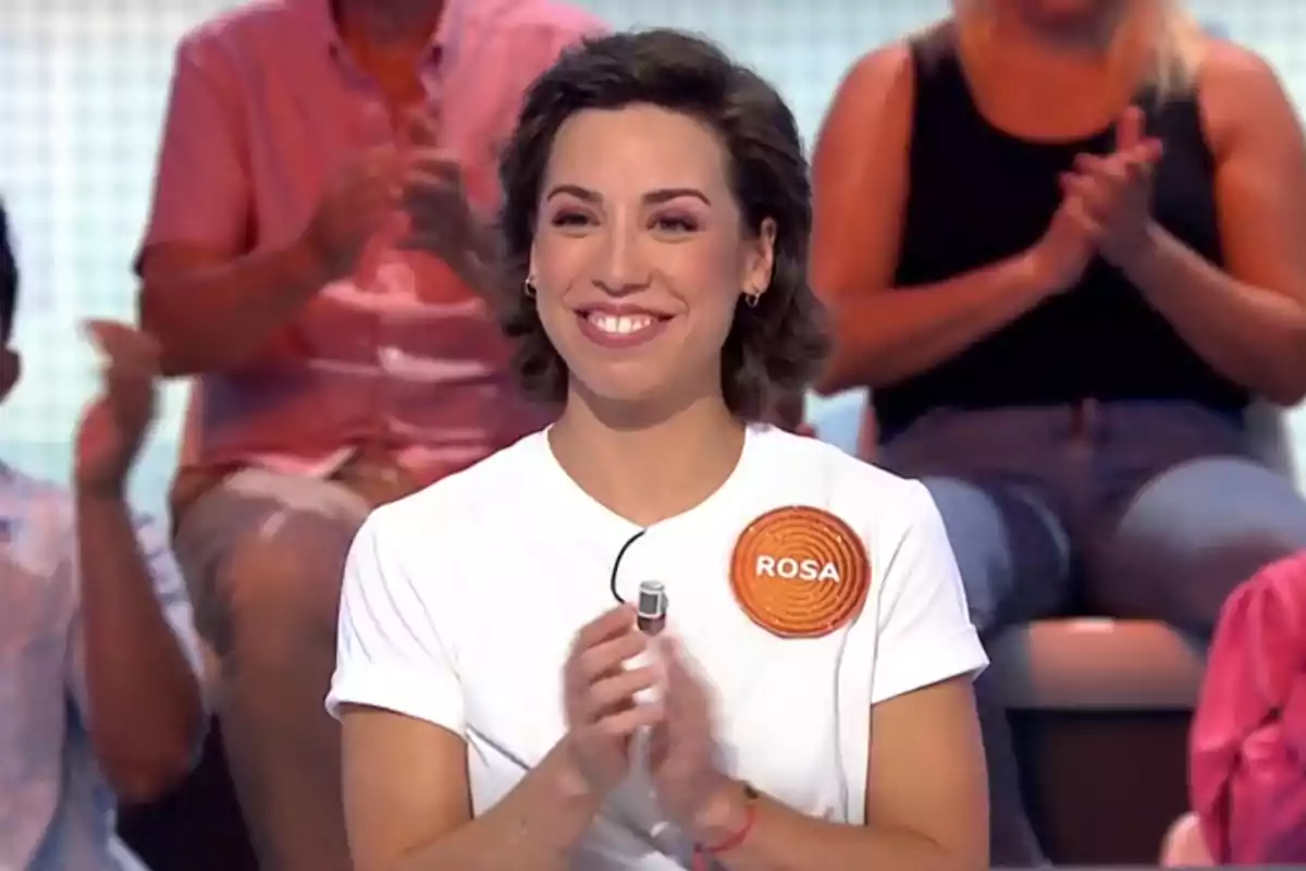 Dona somrient amb samarreta blanca i un distintiu taronja amb el nom de Rosa, aplaudint en un programa de televisió amb públic de fons Dona somrient amb samarreta blanca i un distintiu taronja amb el nom de Rosa, aplaudint en un programa de televisió amb públic de fons