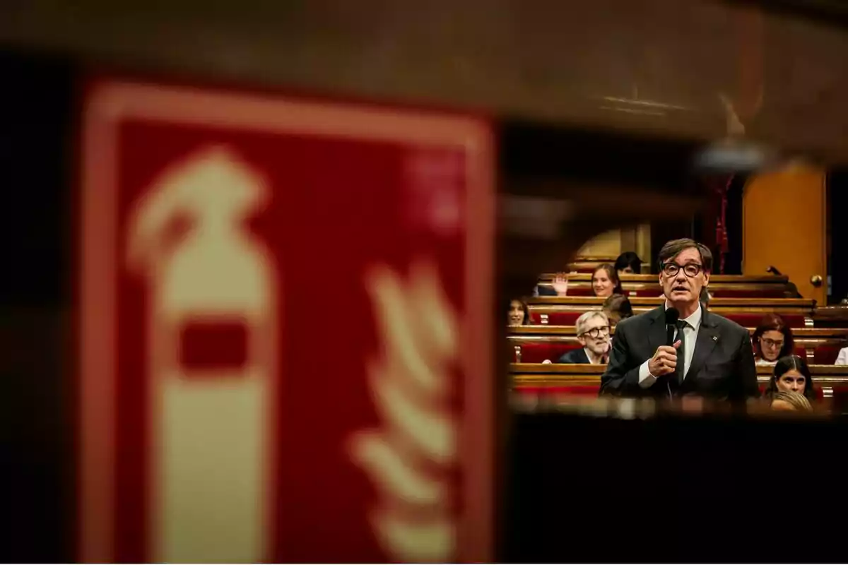 Un home amb vestit i ulleres sosté un micròfon mentre parla en un auditori amb diverses persones assegudes; en primer pla es veu desenfocada una senyal d’extintor. Un home amb vestit i ulleres sosté un micròfon mentre parla en un auditori amb diverses persones assegudes; en primer pla es veu desenfocada una senyal d’extintor.