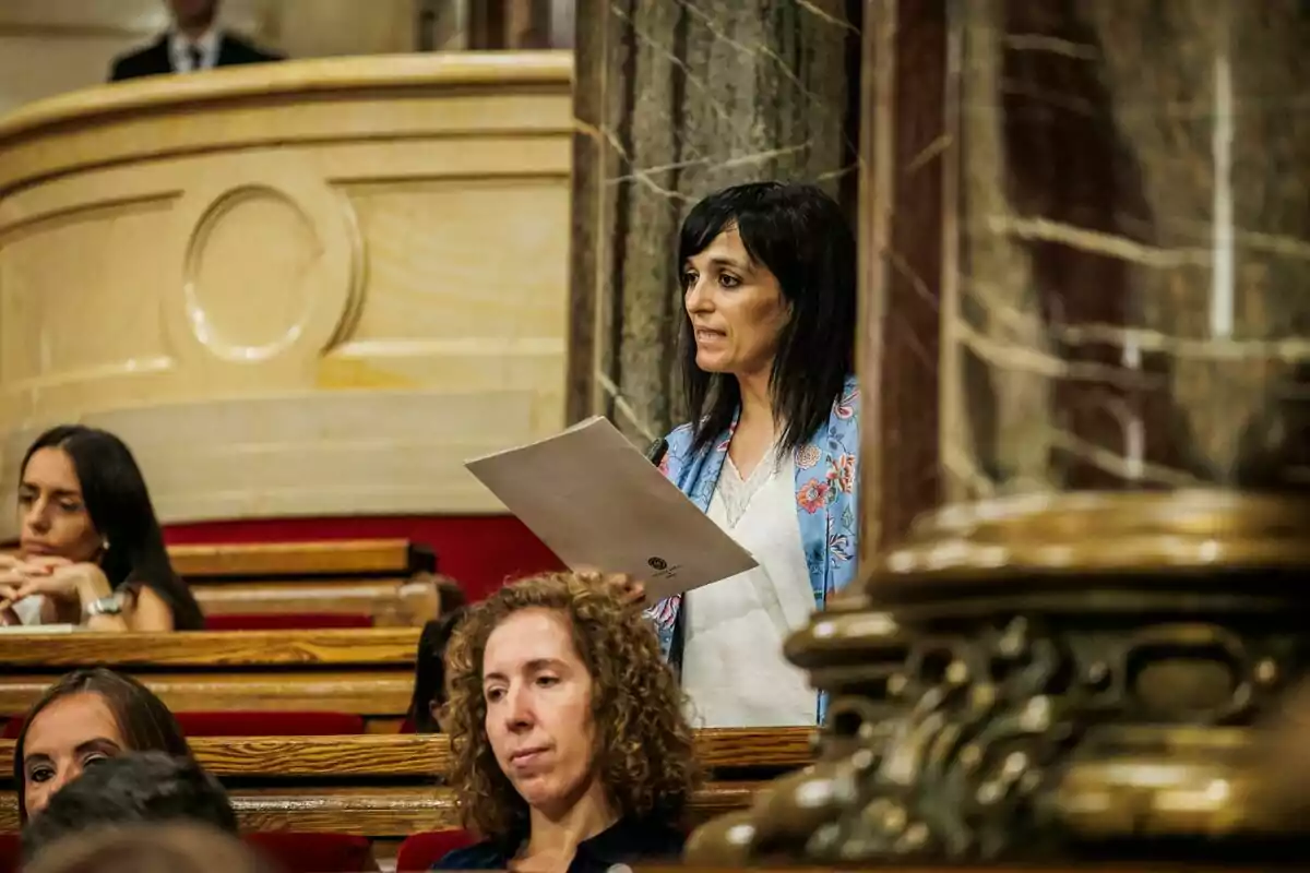 Dona de cabells foscos dreta llegint un document en una sala parlamentària amb altres persones assegudes al voltant Dona de cabells foscos dreta llegint un document en una sala parlamentària amb altres persones assegudes al voltant