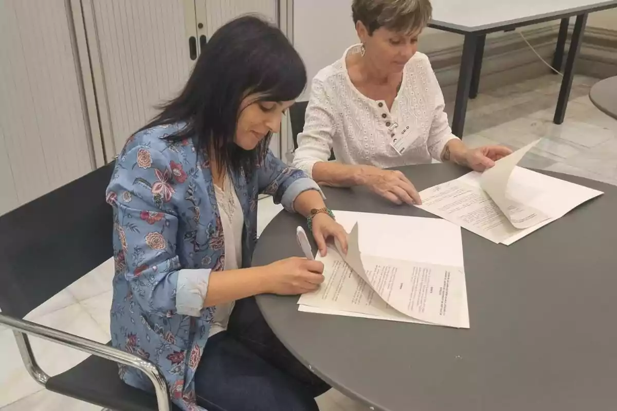 Dues dones assegudes en una taula rodona revisen i signen documents Dues dones assegudes en una taula rodona revisen i signen documents