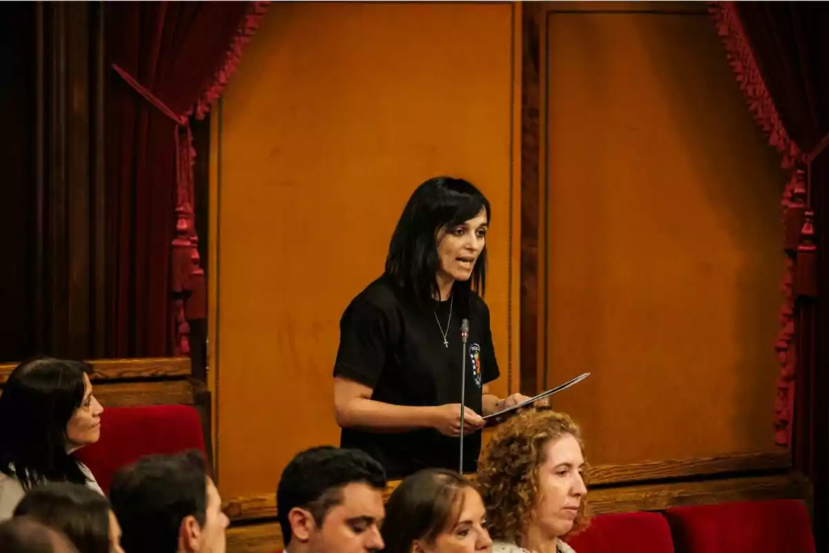 Dona de cabells foscos parlant dreta en un recinte parlamentari amb parets i cortines vermelles mentre sosté uns papers.