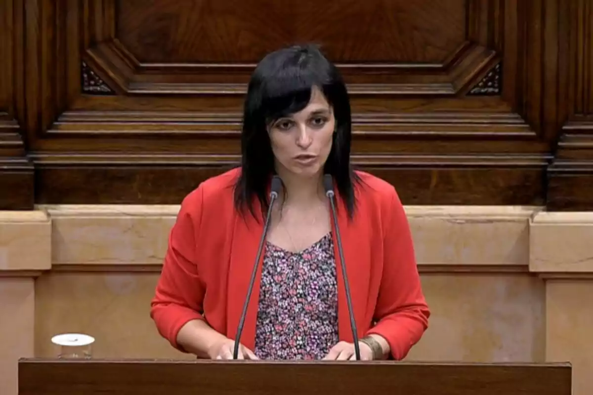Dona de cabell fosc i llis parlant en un faristol amb dos micròfons en un recinte parlamentari, porta jaqueta vermella i brusa de flors