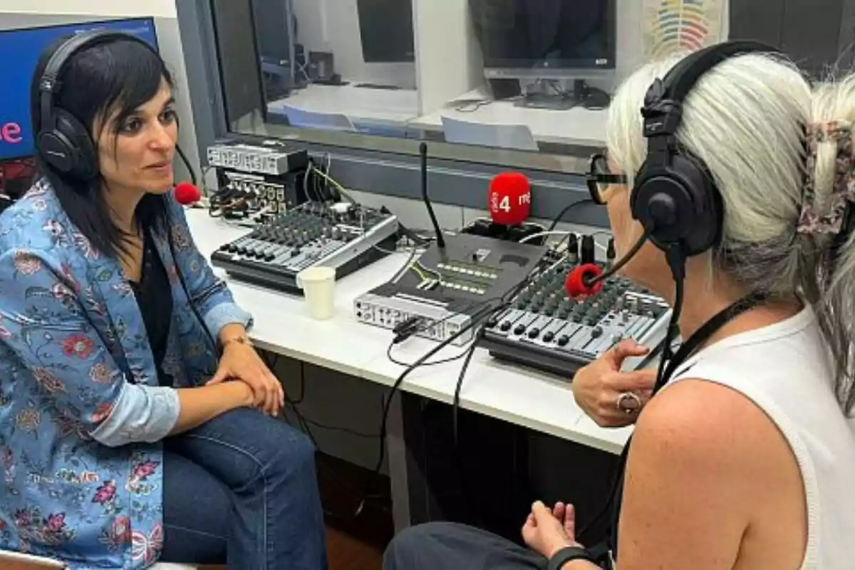 Dues dones amb auriculars i micròfons conversen en una cabina de ràdio envoltades d’equips d’àudio i una taula de mescles.