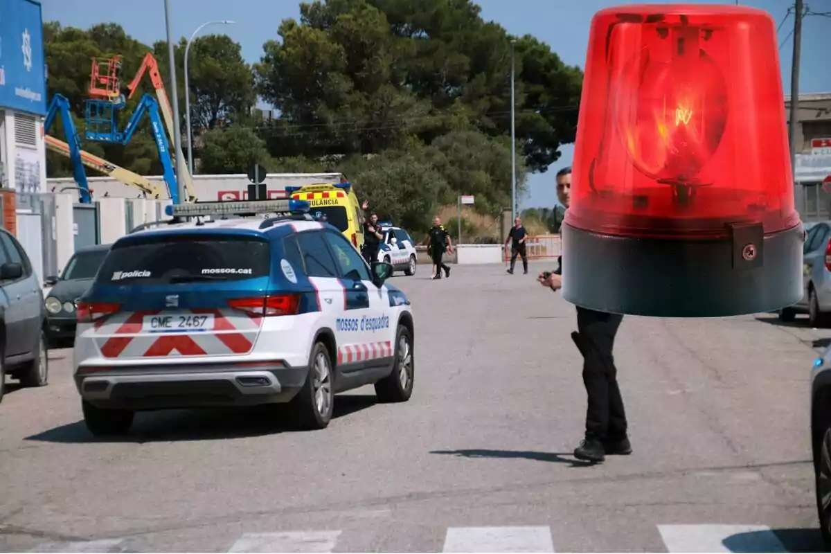 Un cotxe de policia i una ambulància estan estacionats en un carrer amb diversos agents, mentre una sirena vermella de gran mida apareix editada sobre la imatge.