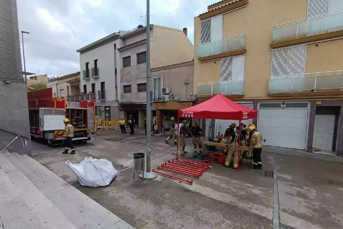 Bombers amb equip d’emergència i un camió treballen en un carrer residencial al costat d’una carpa vermella i eines organitzades a terra.