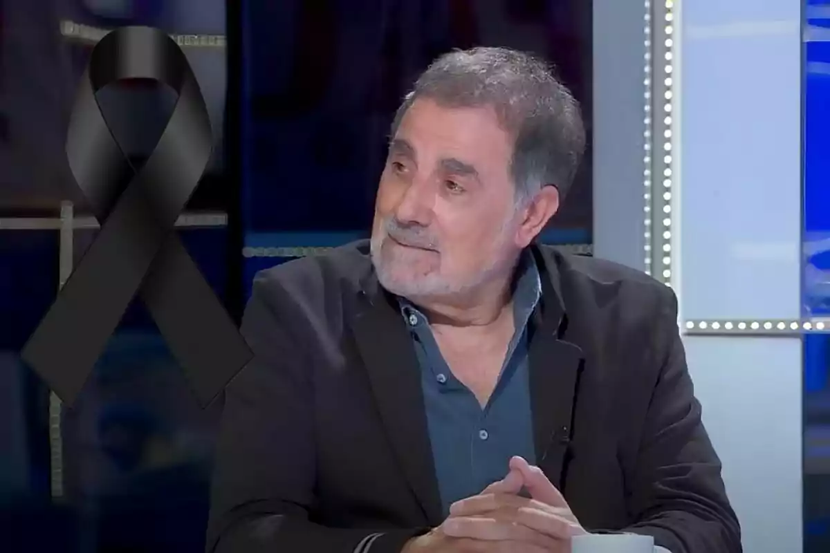 Home de cabells canosos i barba assegut en un estudi de televisió amb un llaç negre de dol al costat esquerre