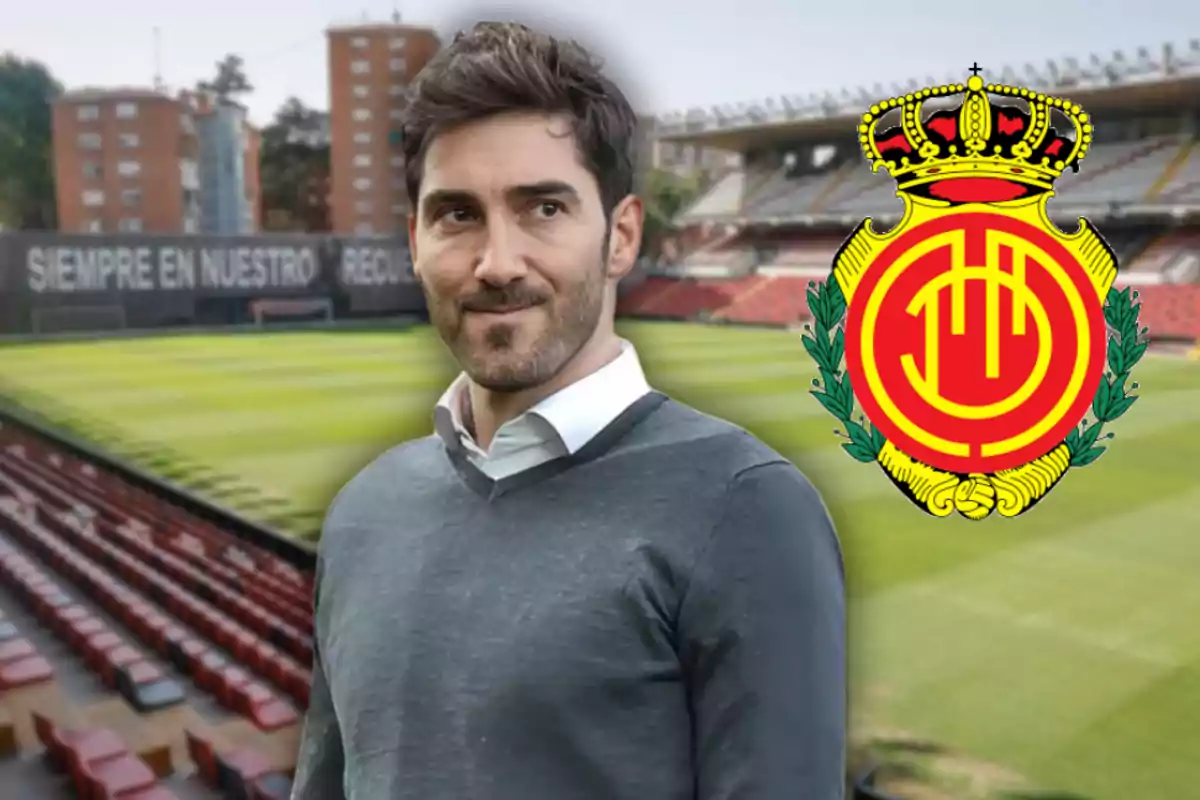 Hombre de cabello oscuro con suéter gris y camisa blanca de pie en un estadio de fútbol con el escudo del Real Mallorca a su lado Hombre de cabello oscuro con suéter gris y camisa blanca de pie en un estadio de fútbol con el escudo del Real Mallorca a su lado