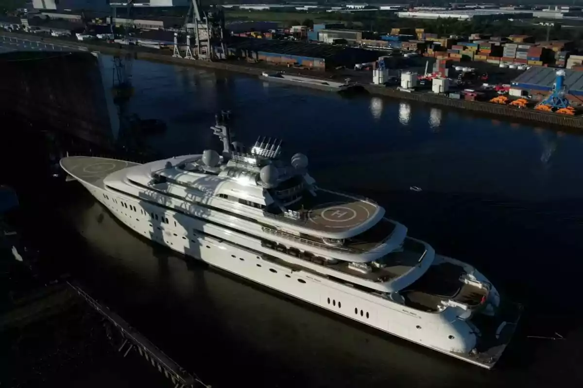 Yate de lujo blanco navegando en un puerto industrial con contenedores y grúas al fondo Yate de lujo blanco navegando en un puerto industrial con contenedores y grúas al fondo