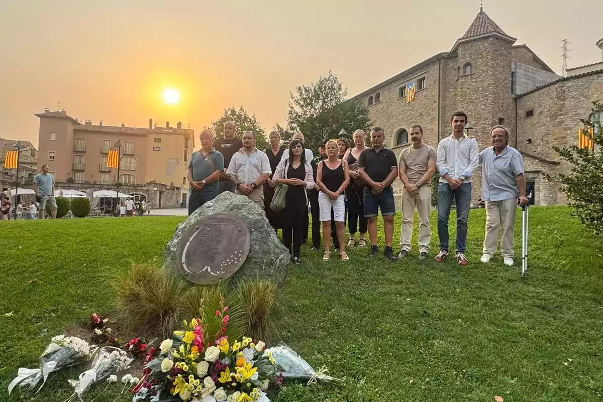 Un grup de persones està reunit en un parc al capvespre davant d’una pedra amb una placa commemorativa i diversos rams de flors, amb edificis antics i banderes catalanes al fons. Un grup de persones està reunit en un parc al capvespre davant d’una pedra amb una placa commemorativa i diversos rams de flors, amb edificis antics i banderes catalanes al fons.