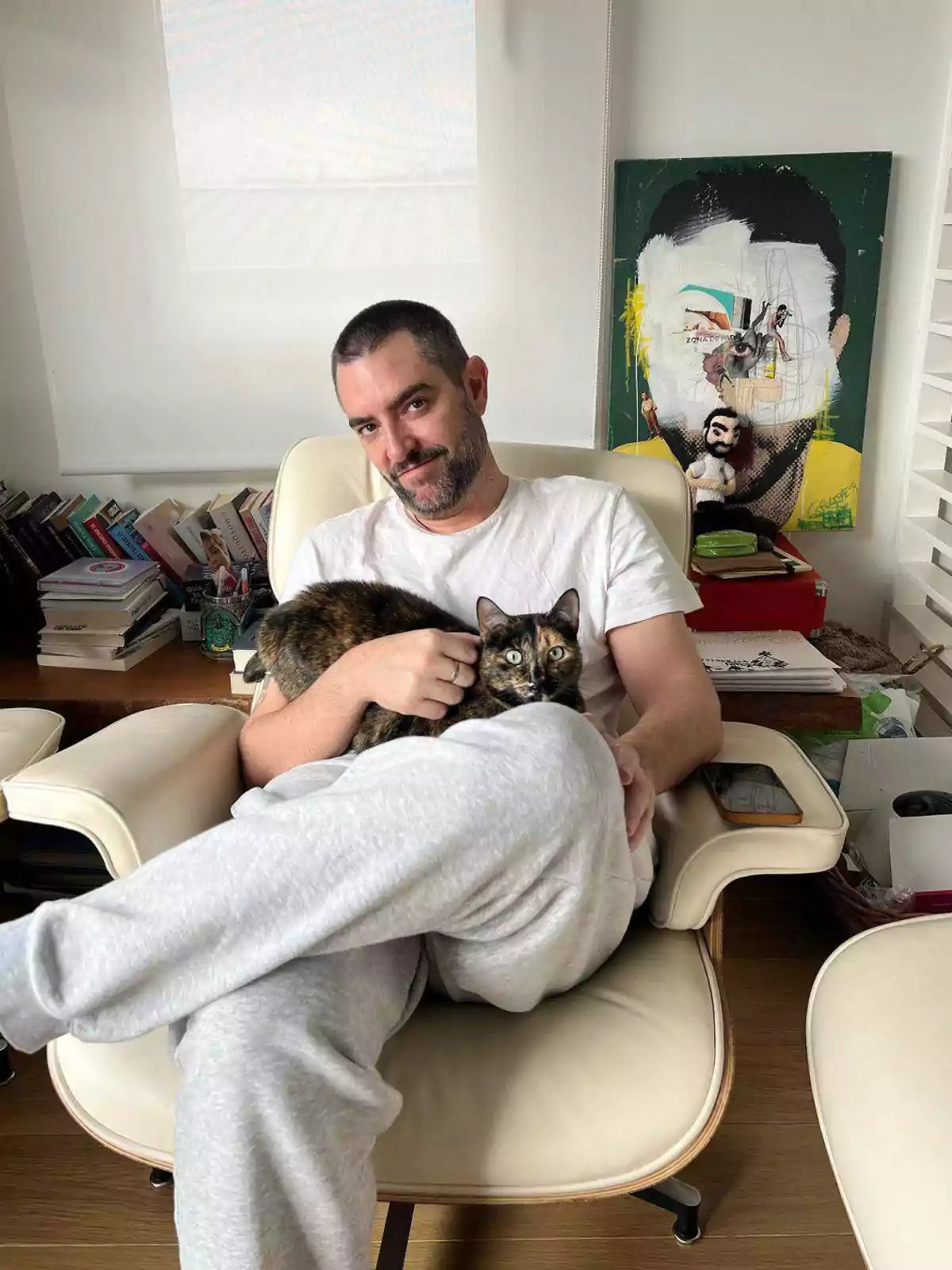 Hombre sentado en un sillón blanco con un gato en brazos en una habitación con libros y una pintura colorida al fondo Hombre sentado en un sillón blanco con un gato en brazos en una habitación con libros y una pintura colorida al fondo
