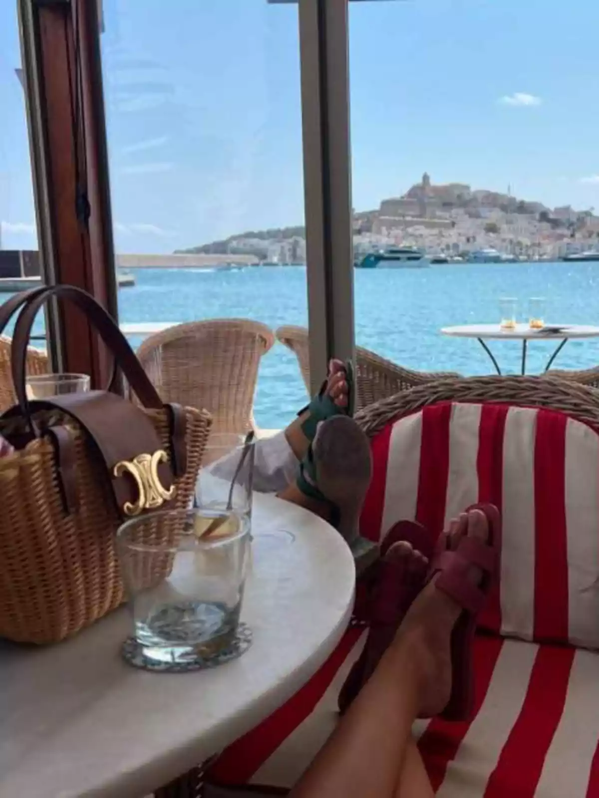 Persona relaxant-se en una terrassa vora el mar amb vista a una ciutat costanera, una taula amb gots i una bossa de vímet. Persona relaxant-se en una terrassa vora el mar amb vista a una ciutat costanera, una taula amb gots i una bossa de vímet.