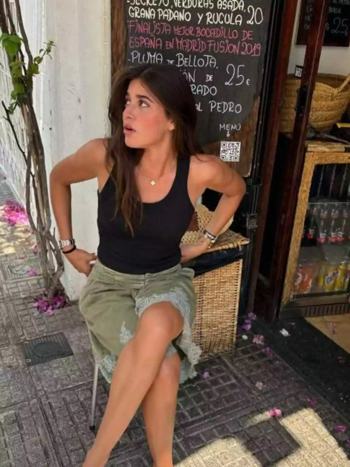 Mujer sentada en una silla al aire libre junto a una pizarra de menú, con expresión de sorpresa y flores en el suelo Mujer sentada en una silla al aire libre junto a una pizarra de menú, con expresión de sorpresa y flores en el suelo