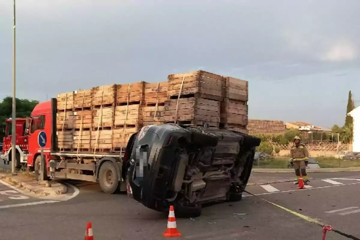 Un camió carregat amb caixes de fusta i un cotxe bolcat en una intersecció mentre un bomber observa l’escena Un camió carregat amb caixes de fusta i un cotxe bolcat en una intersecció mentre un bomber observa l’escena