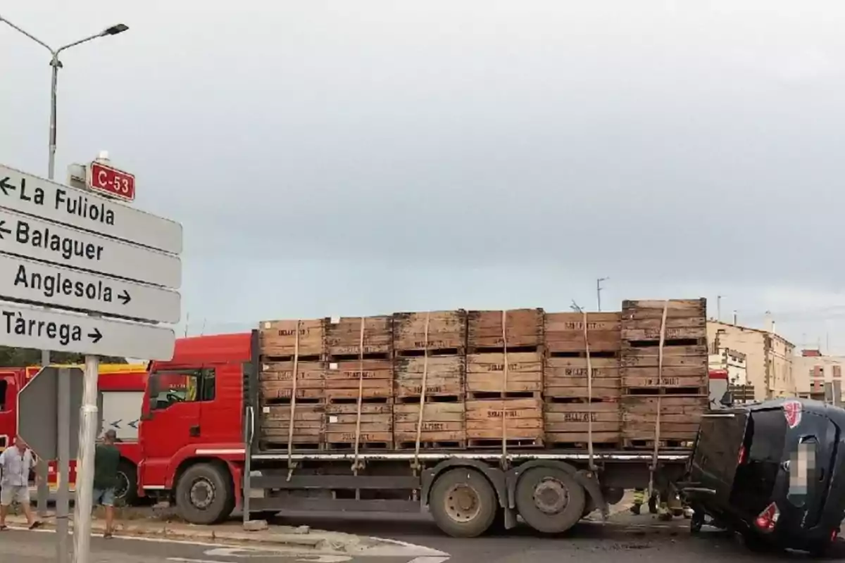Un camió carregat amb caixes de fusta i un cotxe bolcat en una intersecció al costat de senyals de trànsit que indiquen direccions cap a la Fuliola, Balaguer, Anglesola i Tàrrega Un camió carregat amb caixes de fusta i un cotxe bolcat en una intersecció al costat de senyals de trànsit que indiquen direccions cap a la Fuliola, Balaguer, Anglesola i Tàrrega