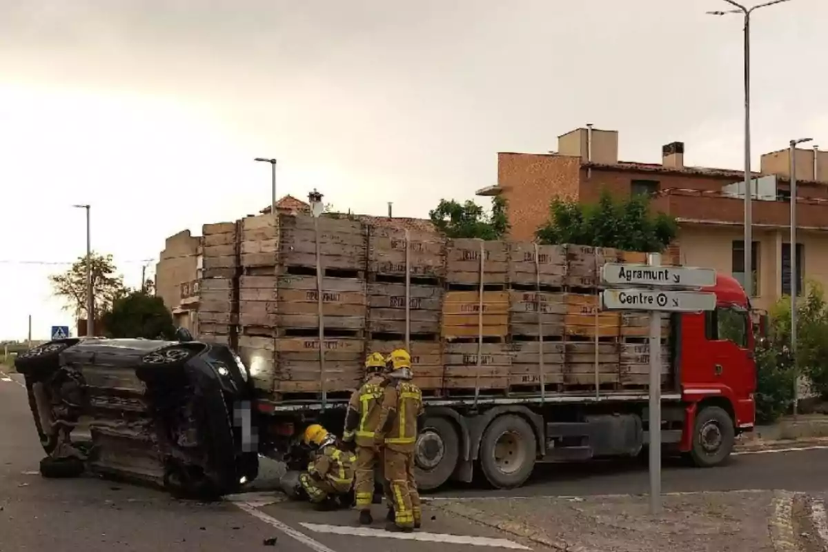 Un camió vermell carregat amb caixes de fusta està aturat en una intersecció al costat d’un cotxe bolcat de costat mentre tres bombers treballen al lloc de l’accident.