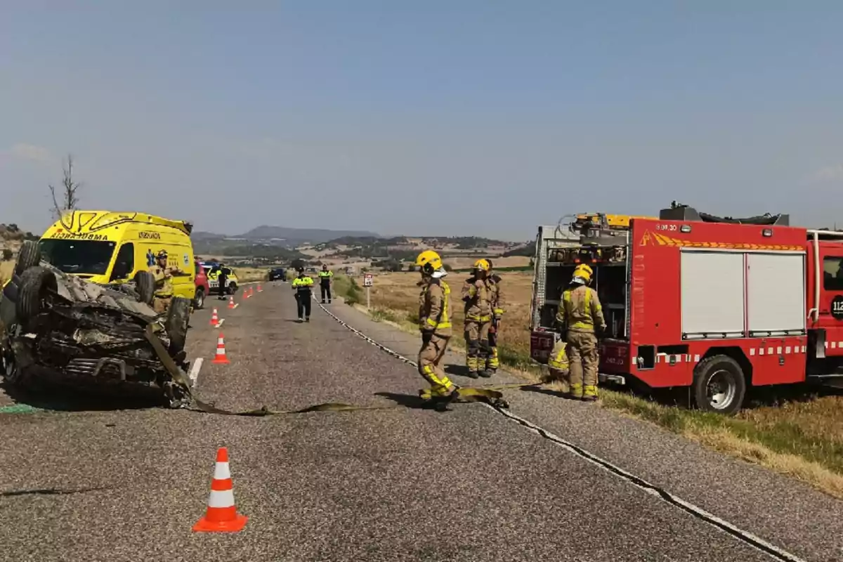 Bomberos y policías atienden un accidente de tráfico en una carretera rural donde un coche está volcado y hay una ambulancia y un camión de bomberos presentes