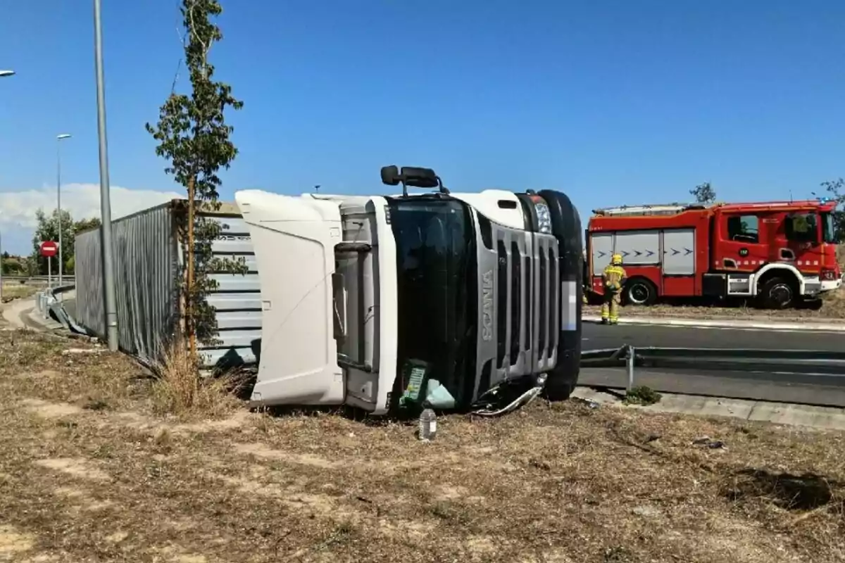 Camión volcado en la orilla de una carretera con un camión de bomberos y un bombero al fondo