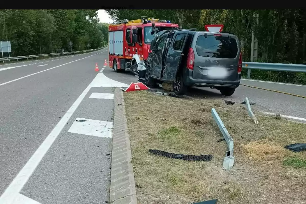 Un coche gris chocado contra una valla de seguridad en una carretera con un camión de bomberos y conos naranjas señalizando el accidente