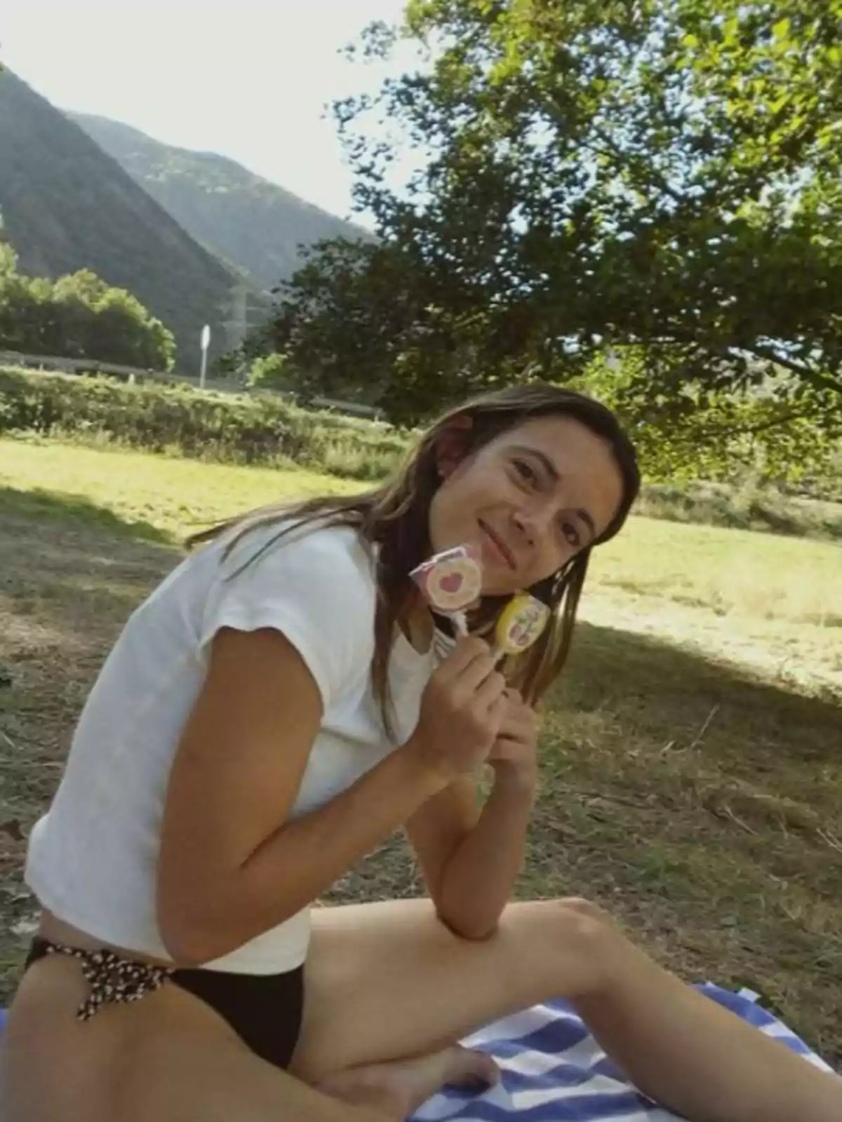 Mujer sentada sobre una manta en un picnic al aire libre sosteniendo dos paletas y sonriendo rodeada de naturaleza y montañas al fondo Mujer sentada sobre una manta en un picnic al aire libre sosteniendo dos paletas y sonriendo rodeada de naturaleza y montañas al fondo
