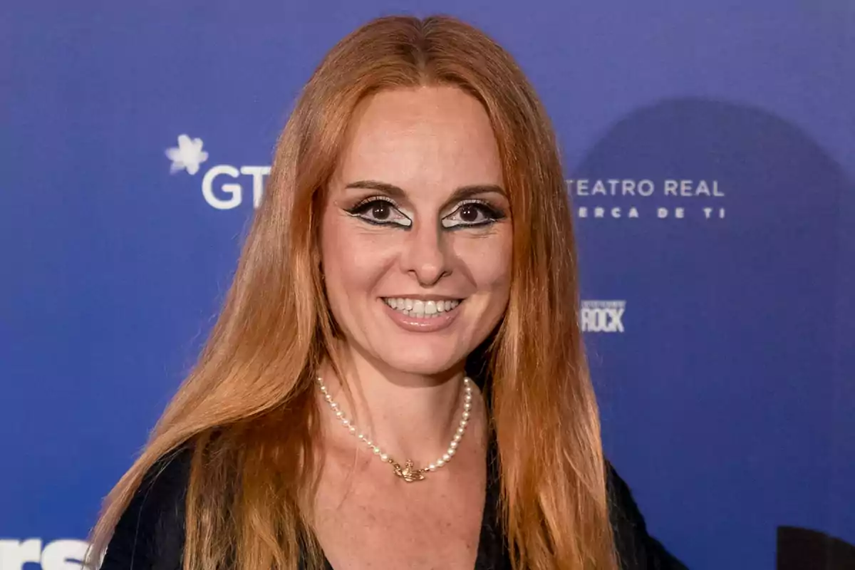 Ana Locking con collar de perlas y maquillaje llamativo en los ojos posando frente a un fondo azul en un evento Ana Locking con collar de perlas y maquillaje llamativo en los ojos posando frente a un fondo azul en un evento