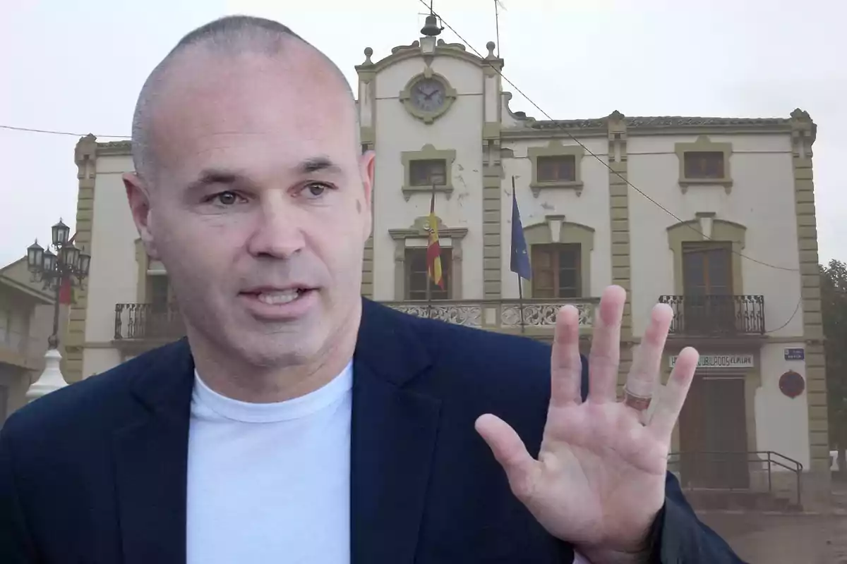 Home calb amb jaqueta fosca i samarreta blanca aixecant la mà davant d’un edifici antic amb banderes i un rellotge a la façana