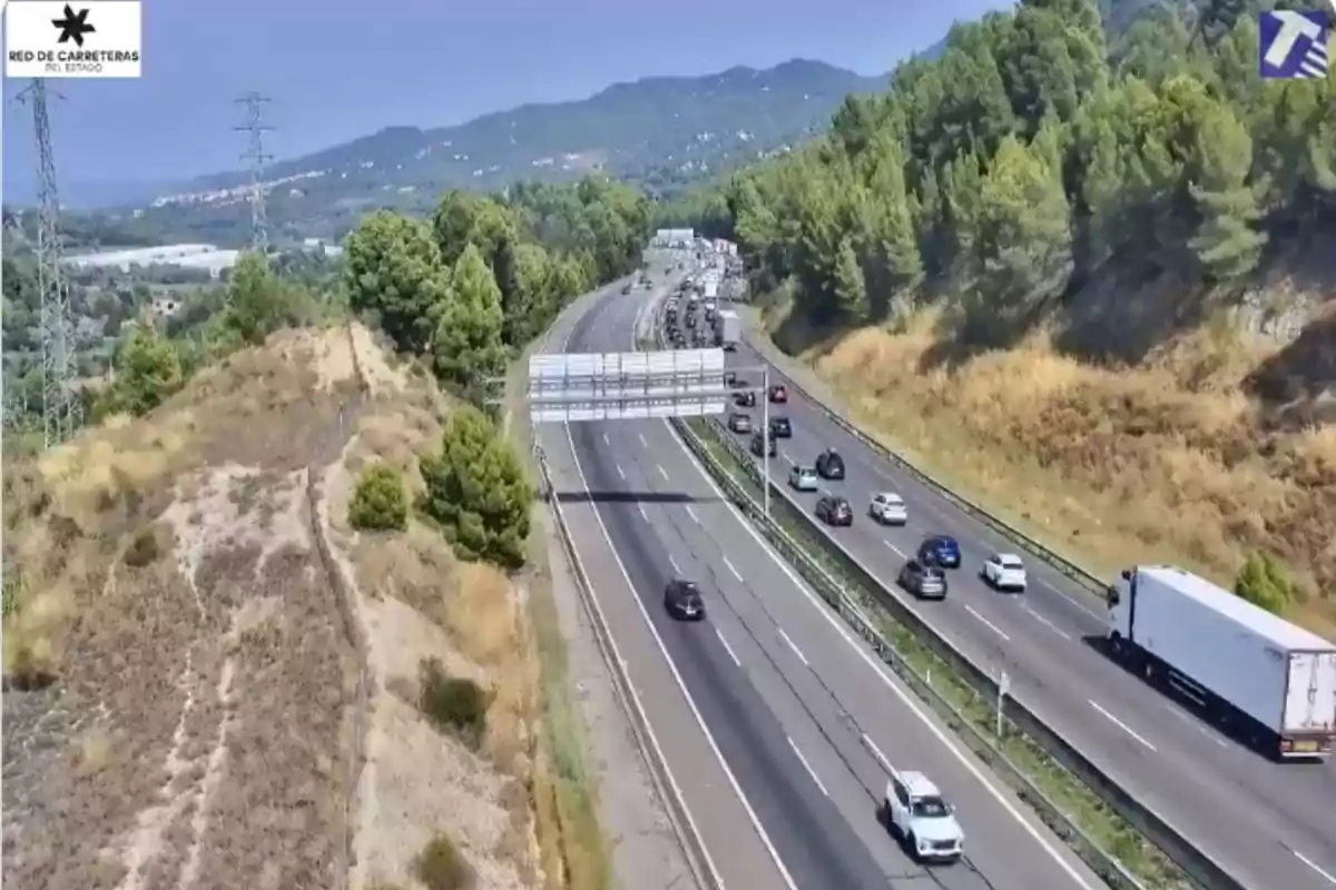 Autopista amb diversos cotxes i un camió circulant envoltada d’arbres i turons sota un cel serè