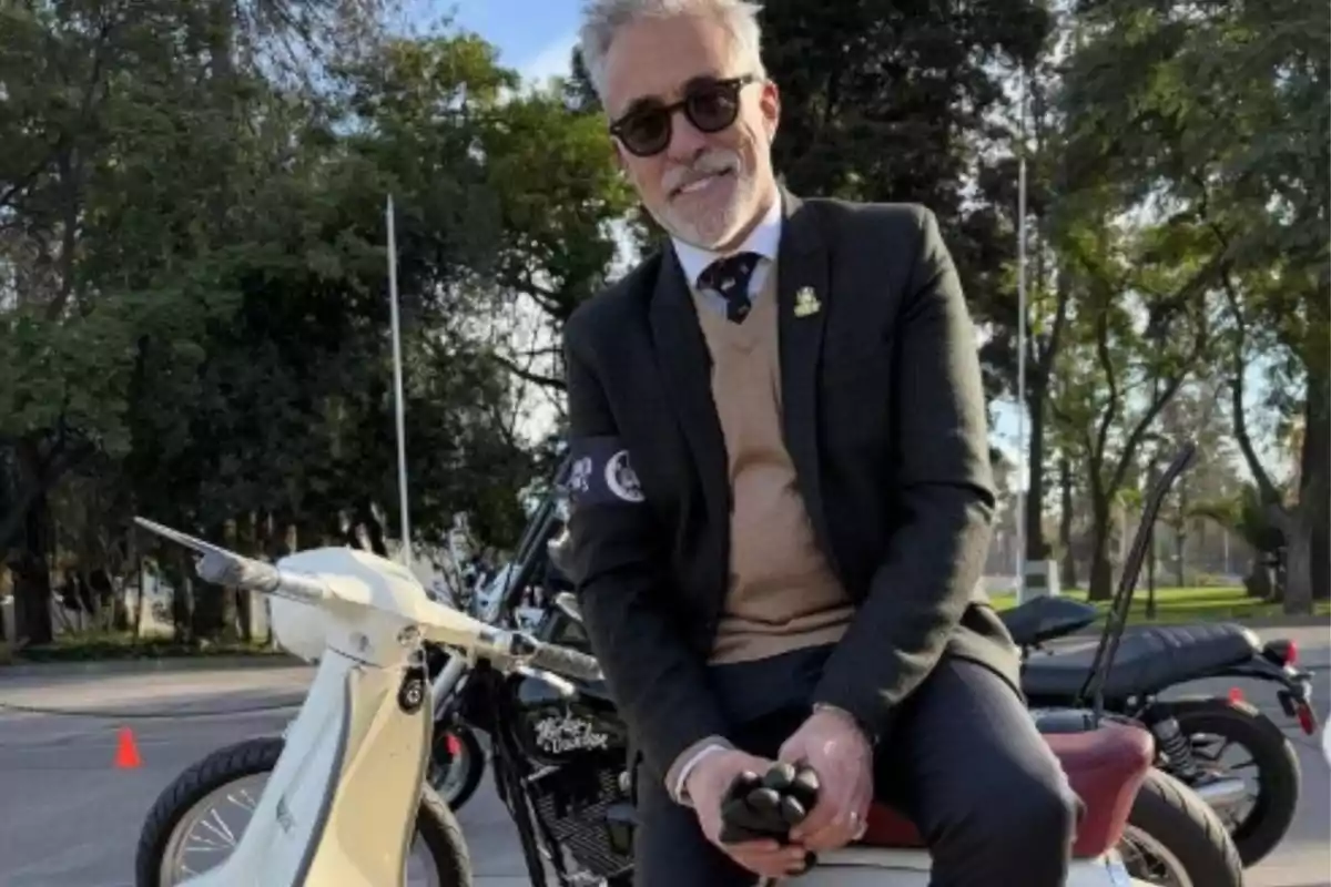Home de cabells canosos i barba assegut en una motocicleta blanca, vestit amb vestit i ulleres de sol, amb arbres i motos al fons Home de cabells canosos i barba assegut en una motocicleta blanca, vestit amb vestit i ulleres de sol, amb arbres i motos al fons