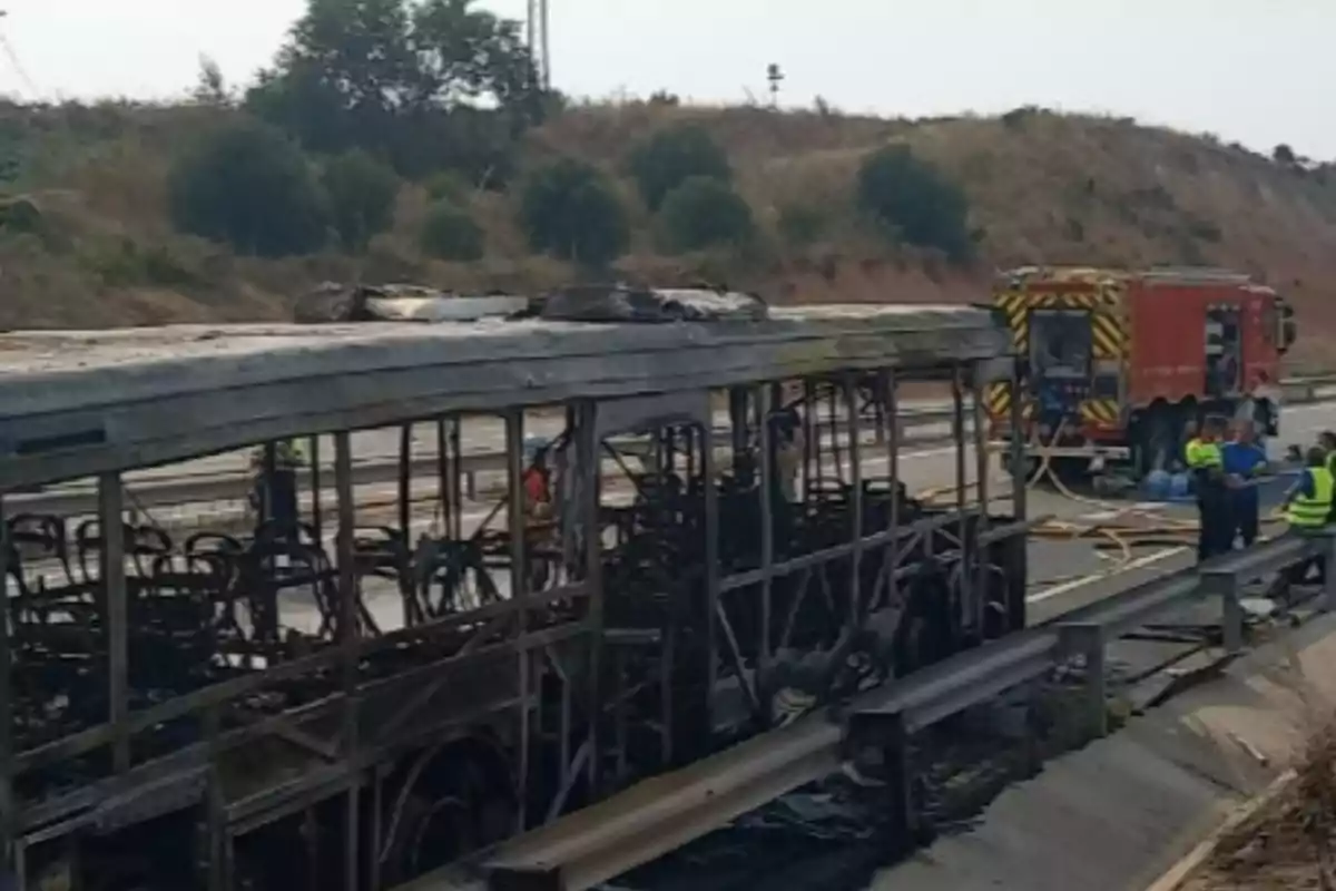 Autobús completament calcinat en una carretera amb bombers i personal d’emergència treballant al lloc. Autobús completament calcinat en una carretera amb bombers i personal d’emergència treballant al lloc.