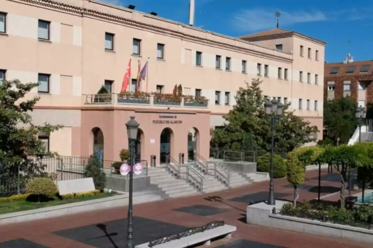 Façana d’un edifici institucional amb diverses finestres, escales d’accés, banderes i una plaça amb bancs i arbres al davant. Façana d’un edifici institucional amb diverses finestres, escales d’accés, banderes i una plaça amb bancs i arbres al davant.