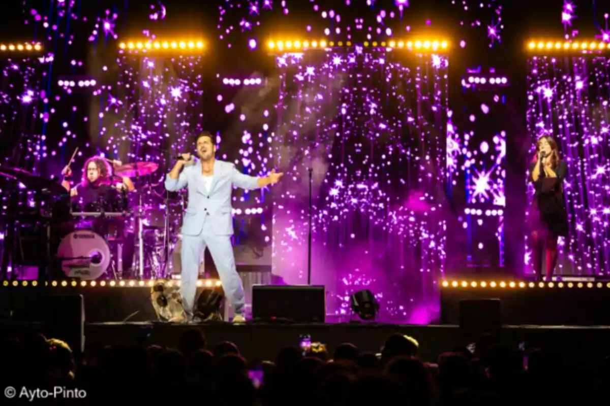 Un cantante con traje claro actúa en el centro del escenario acompañado por una baterista y una corista, con un fondo de luces y efectos morados y amarillos frente a un público. Un cantante con traje claro actúa en el centro del escenario acompañado por una baterista y una corista, con un fondo de luces y efectos morados y amarillos frente a un público.