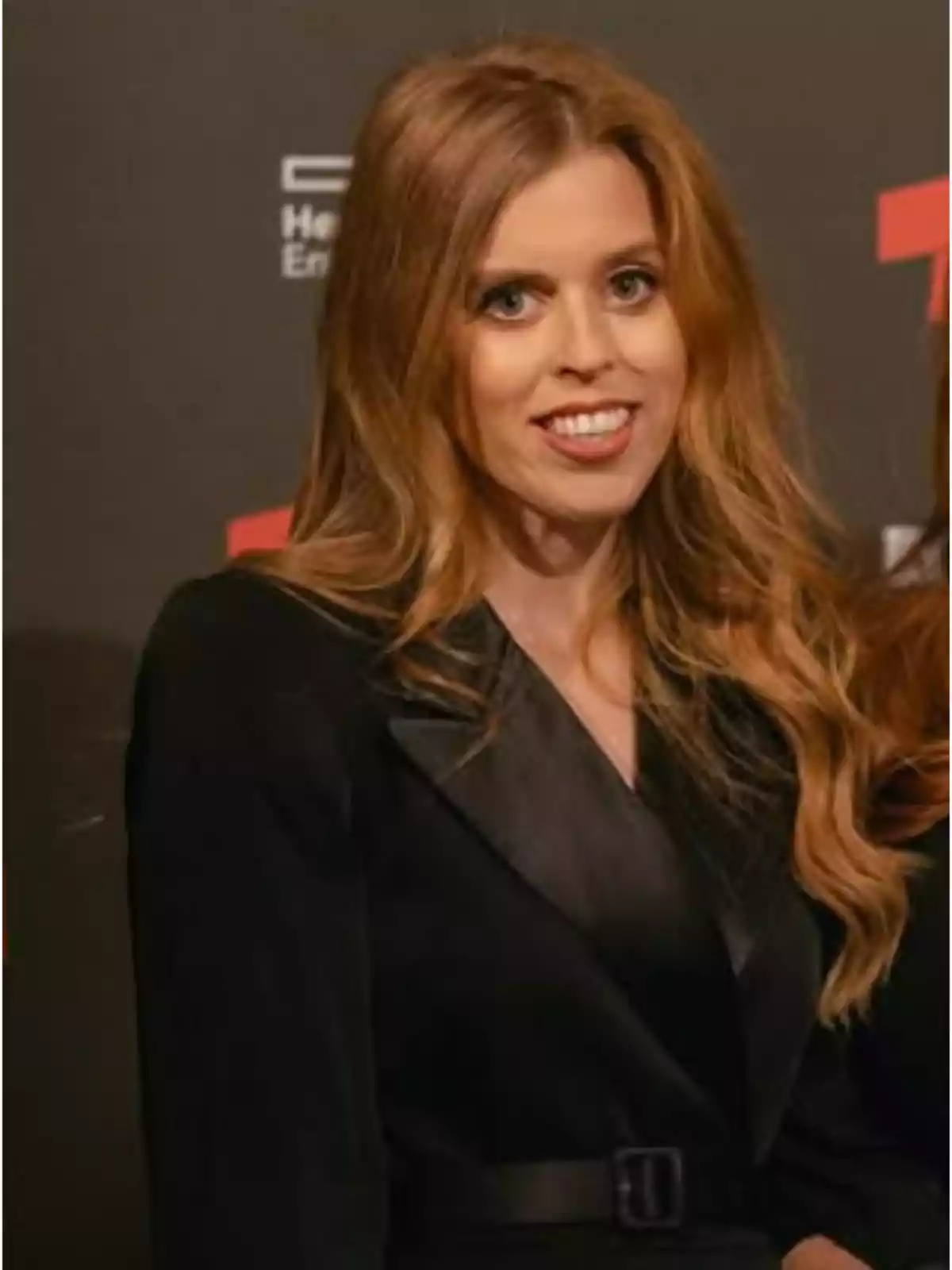 Mujer de cabello largo y rubio con un saco negro posando frente a un fondo oscuro en un evento Mujer de cabello largo y rubio con un saco negro posando frente a un fondo oscuro en un evento