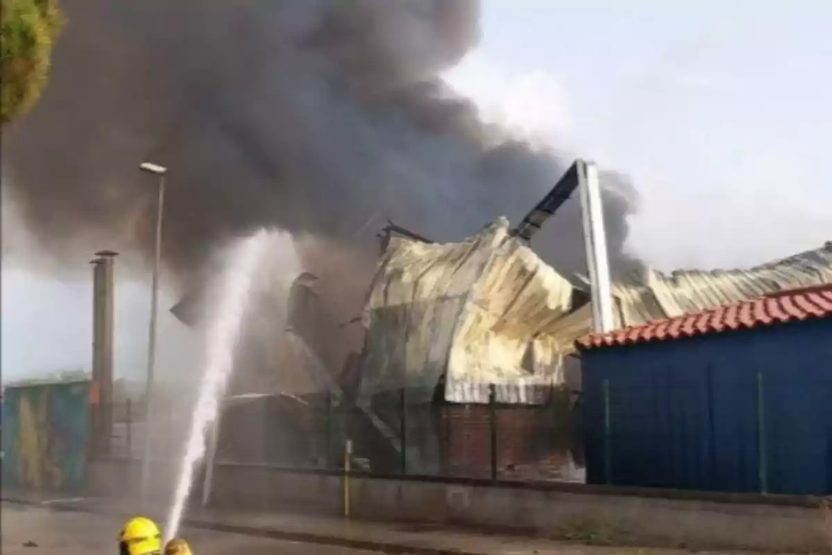 Bomberos apagan un incendio en una nave industrial con una gran columna de humo negro saliendo del edificio. Bomberos apagan un incendio en una nave industrial con una gran columna de humo negro saliendo del edificio.