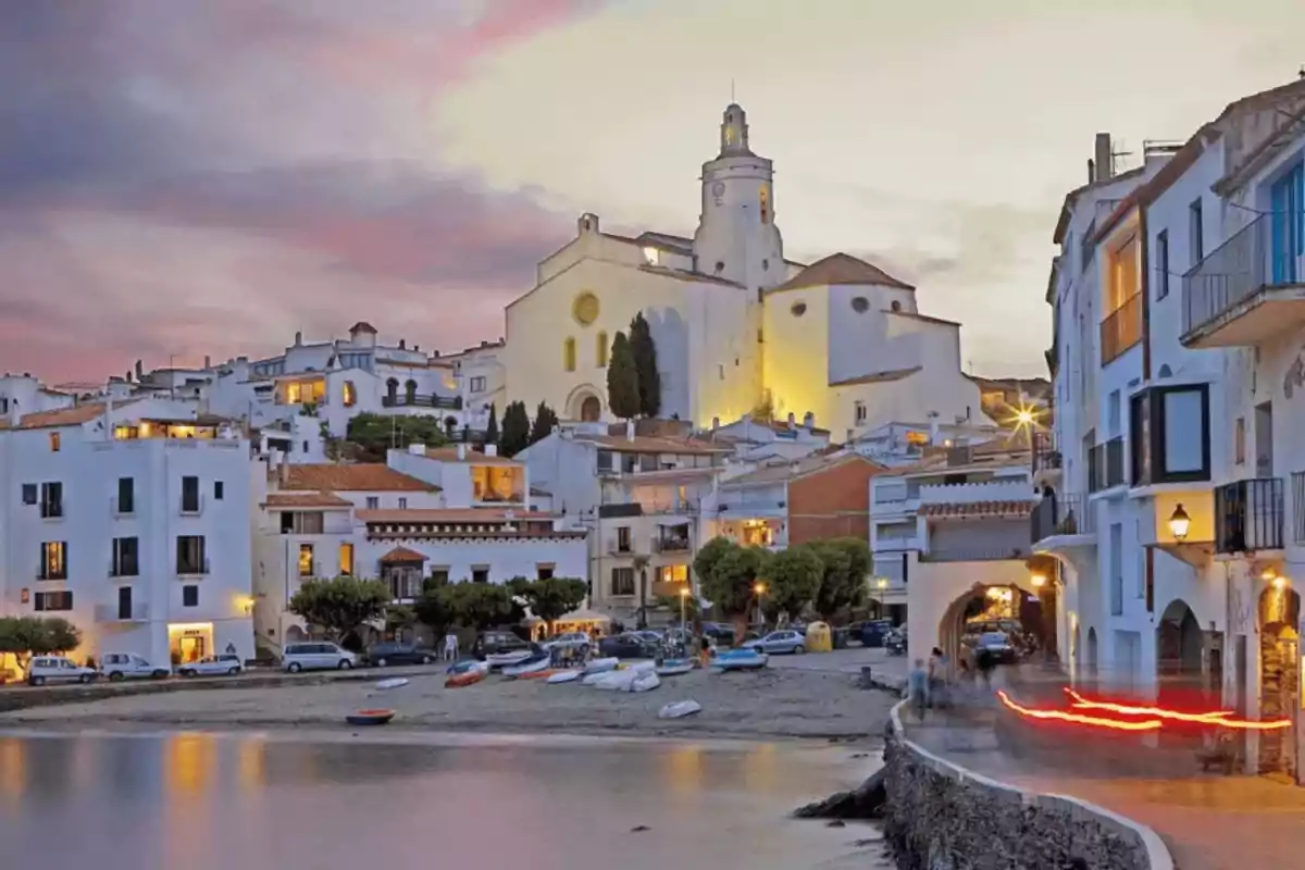 Poble costaner amb cases blanques vora el mar al capvespre i una església il·luminada a la part alta Poble costaner amb cases blanques vora el mar al capvespre i una església il·luminada a la part alta