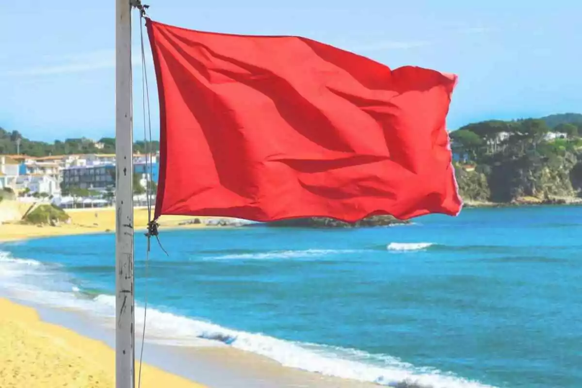 Bandera vermella onejant a la platja amb el mar i edificis al fons