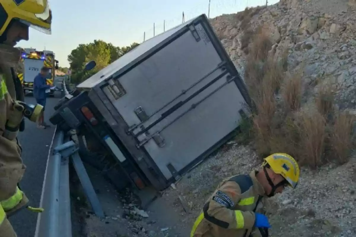 Camión volcado en la cuneta de una carretera con bomberos trabajando en el lugar del accidente.