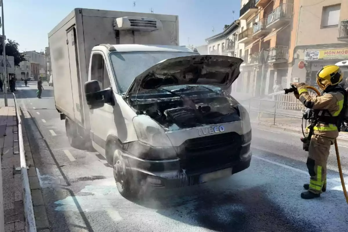 Un bomber apaga l’incendi al motor d’una furgoneta blanca estacionada en un carrer urbà.