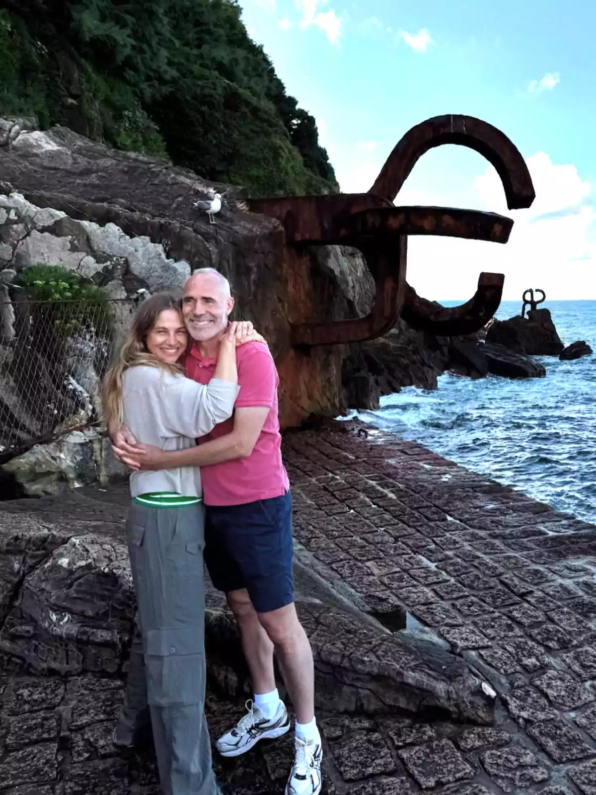 Pareja abrazada sonriente junto al mar con esculturas de hierro en las rocas y vegetación al fondo Pareja abrazada sonriente junto al mar con esculturas de hierro en las rocas y vegetación al fondo