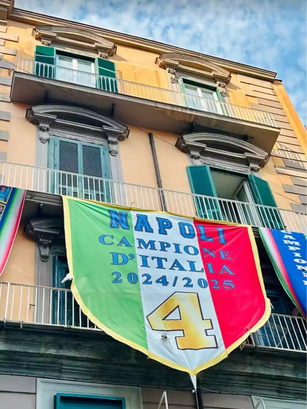 Fachada de un edificio con balcones y ventanas verdes donde cuelga una gran bandera tricolor con el texto Napoli campione d’Italia 2024 2025 y un número 4 amarillo Fachada de un edificio con balcones y ventanas verdes donde cuelga una gran bandera tricolor con el texto Napoli campione d’Italia 2024 2025 y un número 4 amarillo