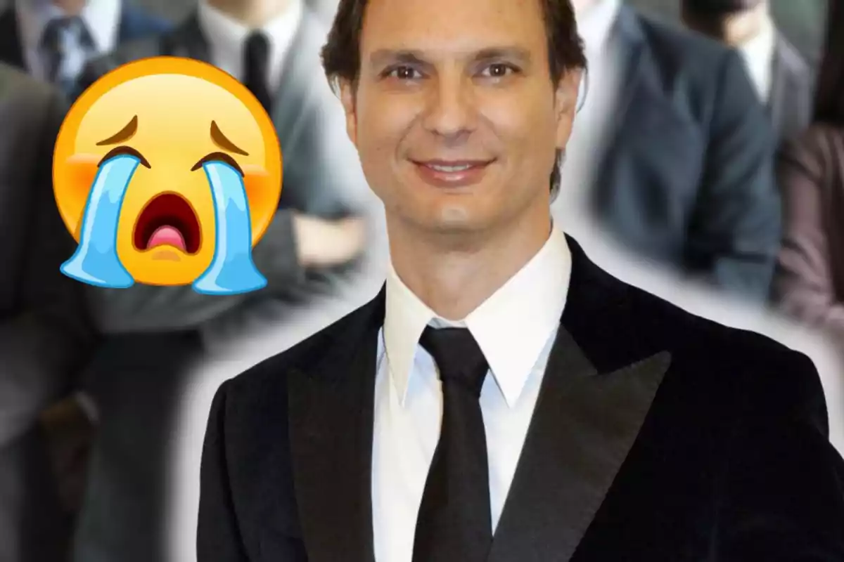 Home amb vestit i corbata somrient amb un emoji de plor gran al seu costat i persones desenfocades al fons