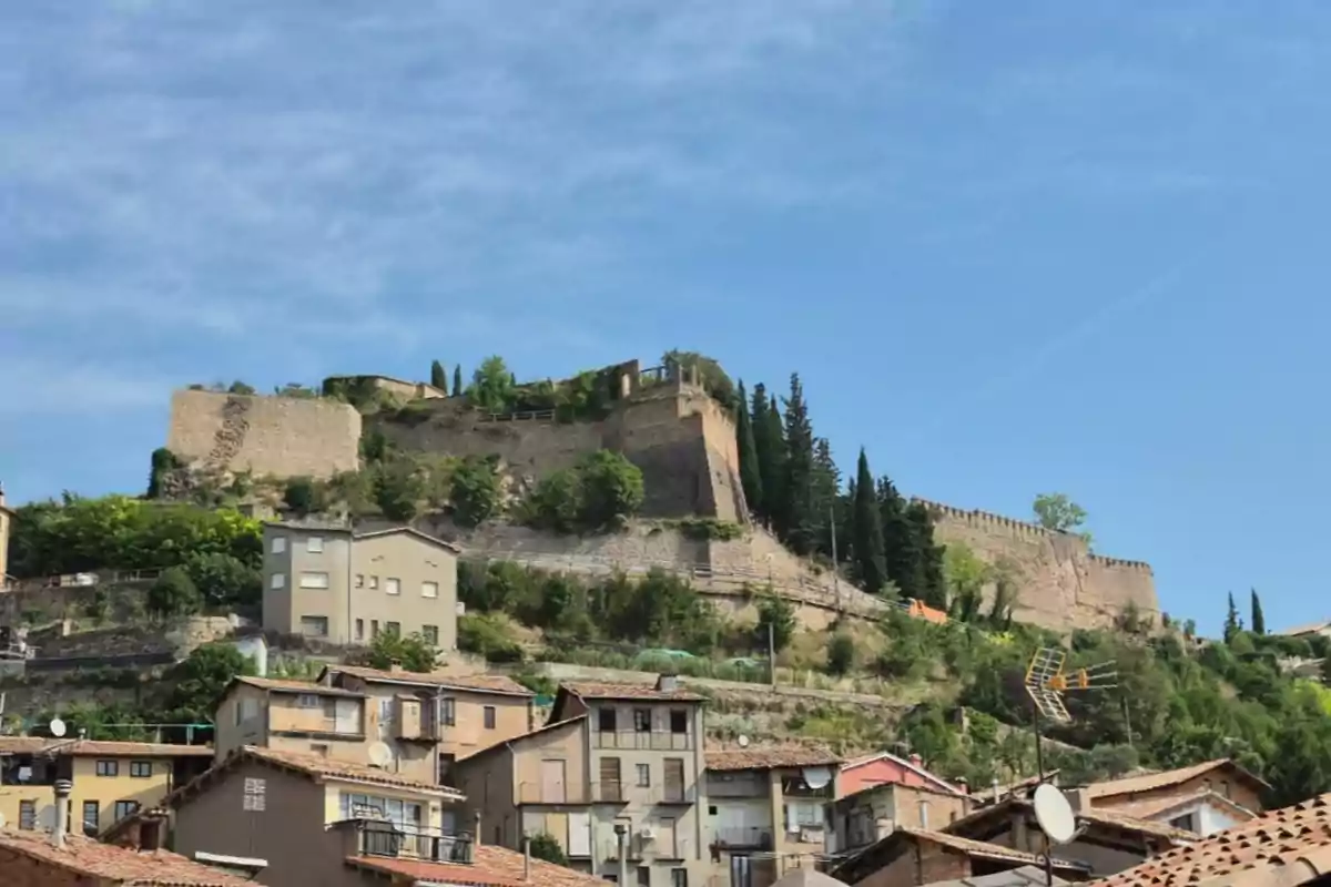 Vista d’una fortalesa de pedra dalt d’un turó envoltada d’arbres i cases a la part baixa sota un cel blau Vista d’una fortalesa de pedra dalt d’un turó envoltada d’arbres i cases a la part baixa sota un cel blau