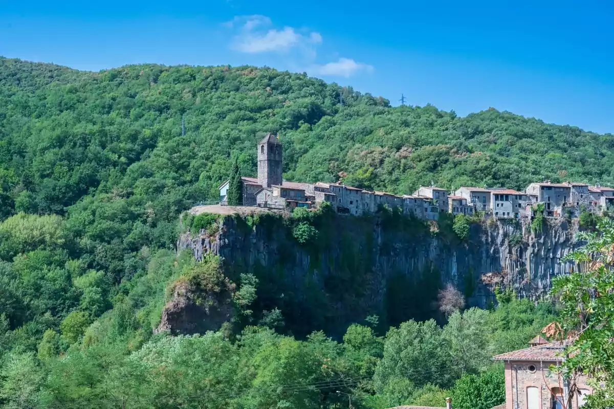 Poble de cases de pedra i una església sobre un penya-segat envoltat de turons verds sota un cel blau Poble de cases de pedra i una església sobre un penya-segat envoltat de turons verds sota un cel blau