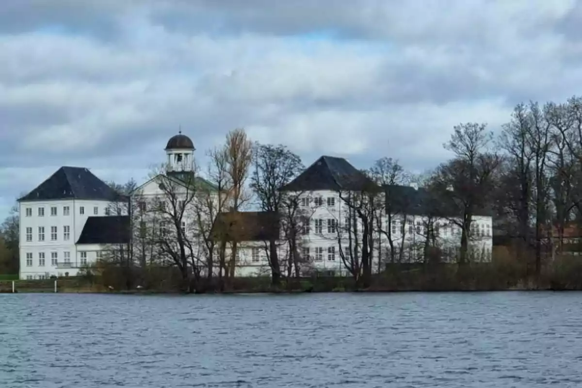 Edifici gran de color blanc amb teulada fosca al costat d’un llac i envoltat d’arbres sense fulles sota un cel ennuvolat. Edifici gran de color blanc amb teulada fosca al costat d’un llac i envoltat d’arbres sense fulles sota un cel ennuvolat.