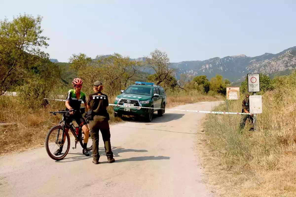 Un ciclista és detingut per una guarda forestal en un camí rural bloquejat amb cinta, mentre un vehicle oficial està estacionat i un altre guarda forestal assegura la zona, amb muntanyes al fons. Un ciclista és detingut per una guarda forestal en un camí rural bloquejat amb cinta, mentre un vehicle oficial està estacionat i un altre guarda forestal assegura la zona, amb muntanyes al fons.