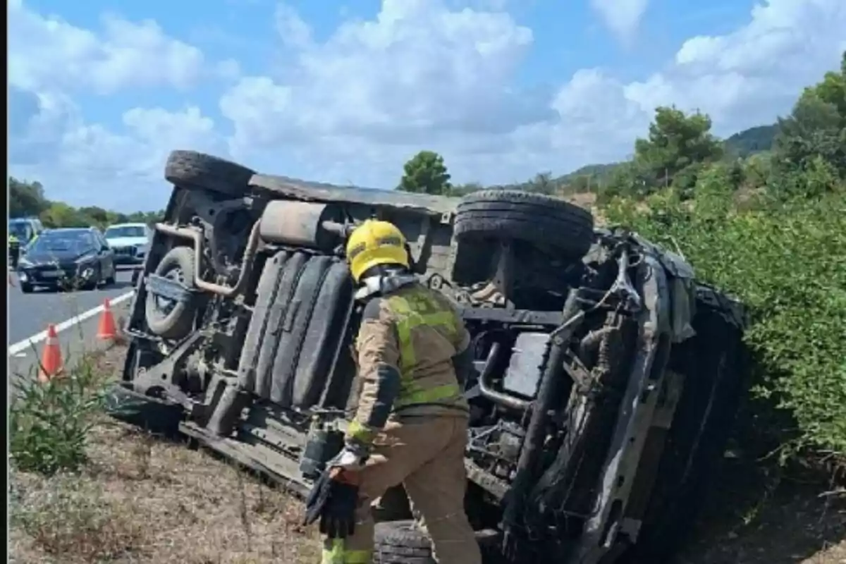 Un bomber amb casc groc inspecciona un camió bolcat de costat a la vora d’una carretera mentre diversos cotxes passen i hi ha cons taronges que senyalitzen l’accident.