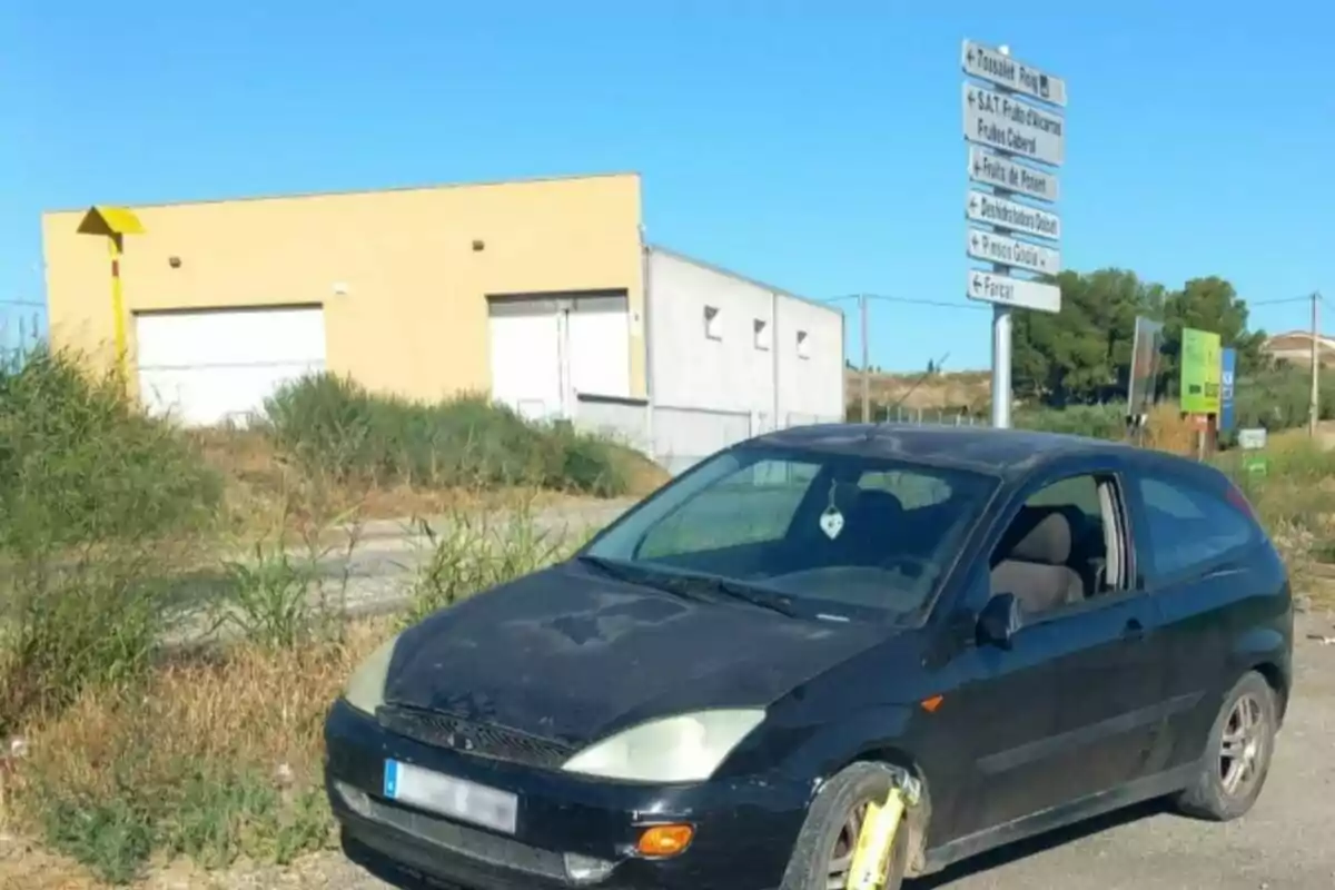 Un cotxe negre amb una trampa per a rodes en una zona rural al costat d’un edifici groc i senyals de trànsit