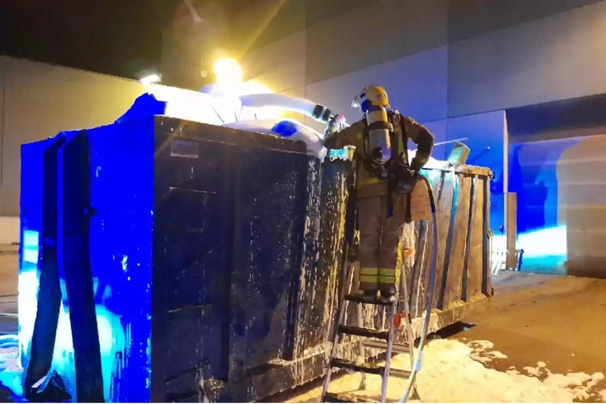 Bombero con equipo de protección apaga un incendio en un contenedor industrial usando espuma mientras está de pie en una escalera