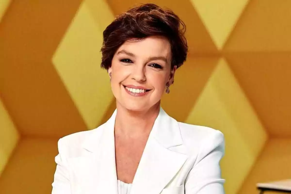 Mujer de cabello corto y castaño sonriendo, vestida con saco blanco y fondo geométrico amarillo Mujer de cabello corto y castaño sonriendo, vestida con saco blanco y fondo geométrico amarillo